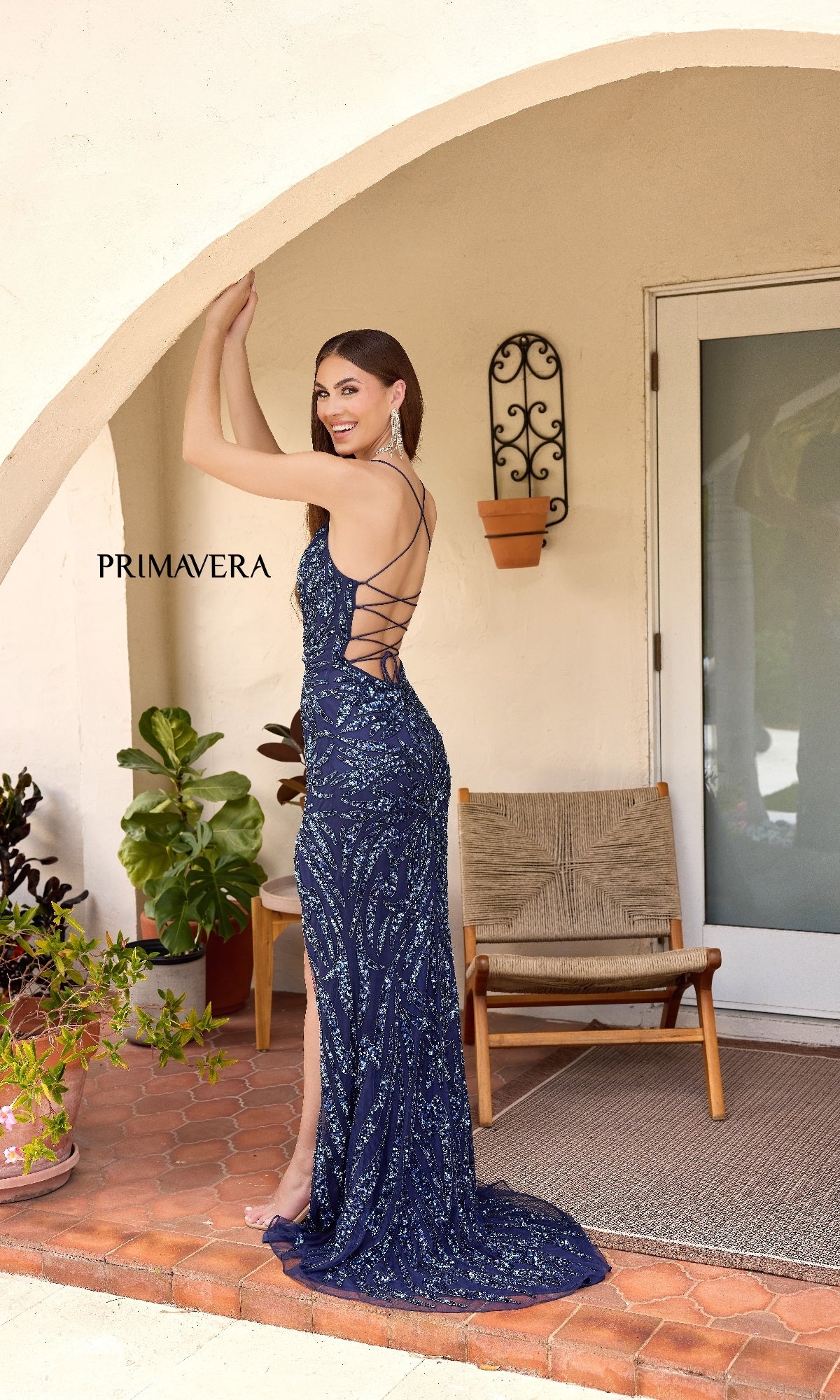 A woman poses on a tiled patio in the Primavera 4303, a navy blue, backless long sequin prom dress—an eye-catching, elegant choice for any formal occasion.